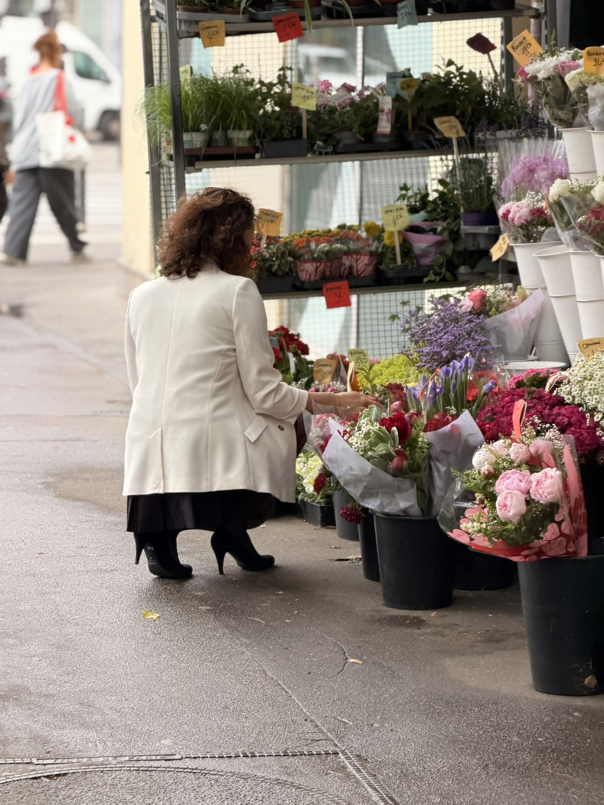 Lena im hellen Mantel kniet vor einem Blumenstand auf der Strasse und sucht Blumen aus