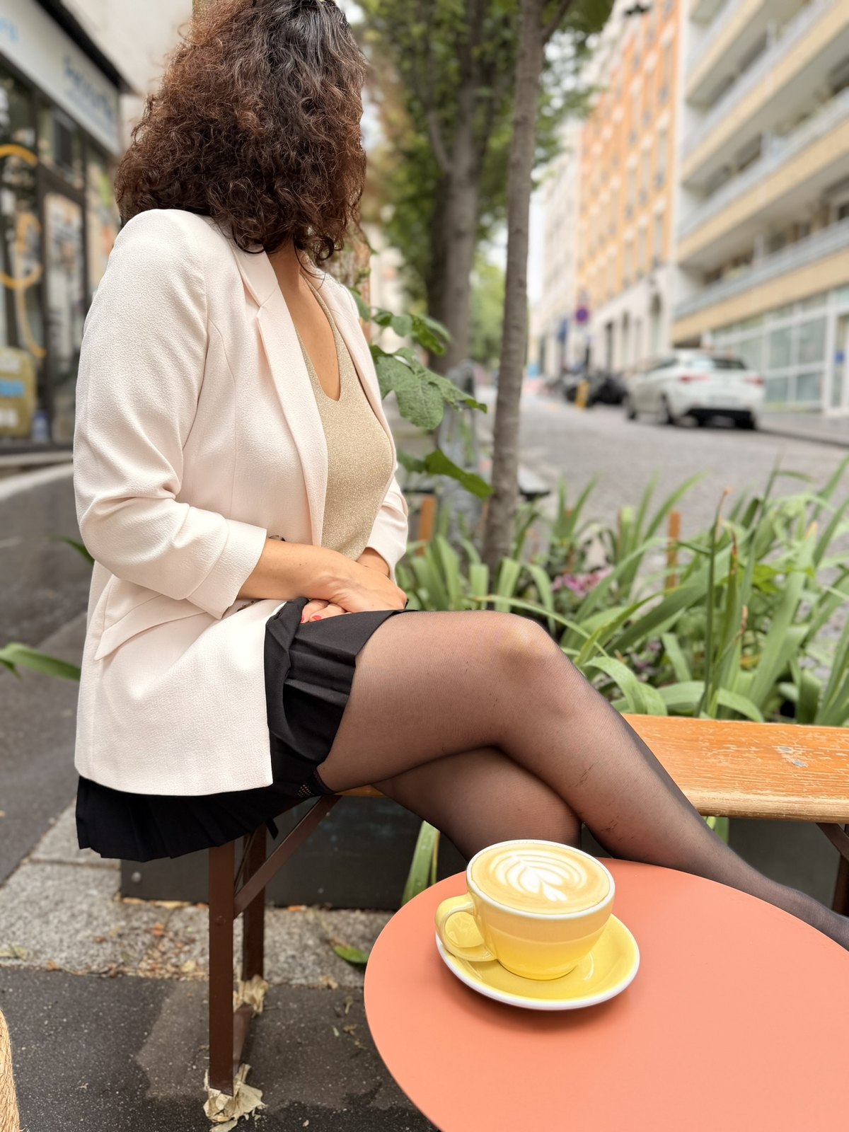 Lena sitzt im hellen Blazer auf einer Caféterrasse, vor ihr ein Cappuccino auf einem runden Tisch