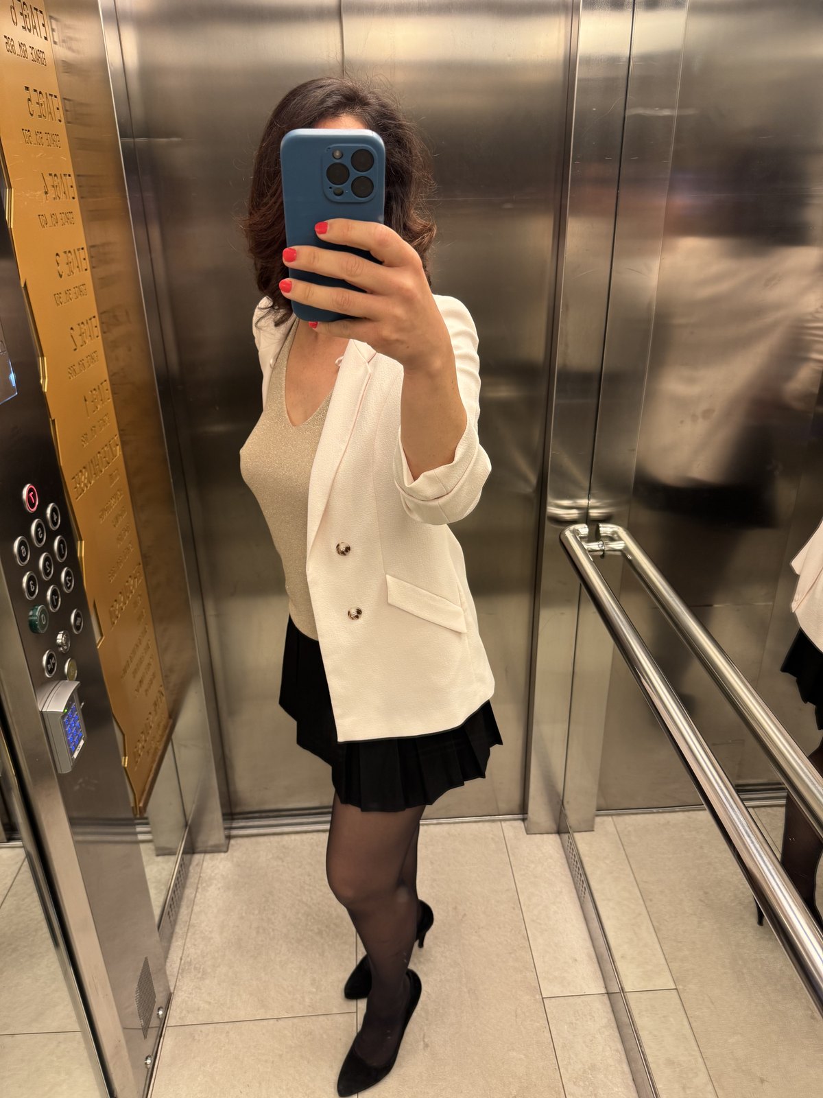 Lena macht ein Spiegel-Selfie im Lift, weisser Blazer, schwarzer Faltenrock, das Natel verdeckt ihr Gesicht